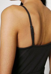 Černé bezramínkové šaty s přiléhavým tělem, vybavené tenkými nastavitelnými ramínky a hladkým, texturovaným materiálem. Zadní výstřih je rovný.