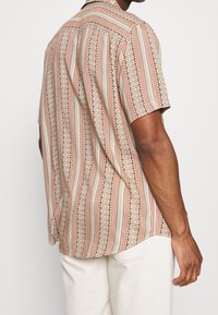 Chemise à manches courtes beige avec des motifs géométriques rouges, bruns et blancs. Confectionnée en tissu léger avec une coupe décontractée et une texture subtile.