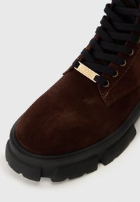 Stivale in suede marrone scuro con lacci neri spessi e suola nera chunky, caratterizzato da un piccolo tag dorato inciso con "Steve Madden" sui lacci.