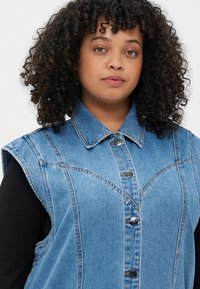 Svetlo modra denim brezrokavnica s poudarjenim ovratnikom, sprednjim gumbom in poševnimi šivi. Teksturni material s sproščenim dizajnom.