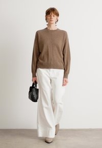 Καφέ πλεκτή ζακέτα με μακριά μανίκια και στρογγυλή λαιμόκοψη, συνδυασμένη με λευκά παντελόνια σε φαρδιά γραμμή. Κρατάει μια μικρή μαύρη τσάντα με πλεκτό σχέδιο.