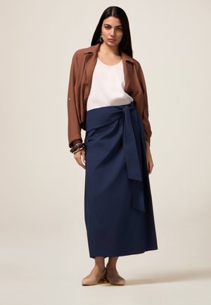 Bruine oversized jas, witte top en marineblauwe wikkelrok met een strik. De outfit heeft een gladde structuur en een eenvoudig, minimalistisch ontwerp.