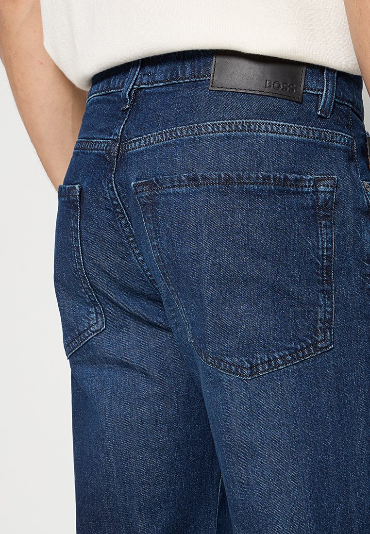 Jean en denim bleu foncé avec un design classique à cinq poches, présentant une étiquette en cuir et des détails de couture visibles. Texture lisse.