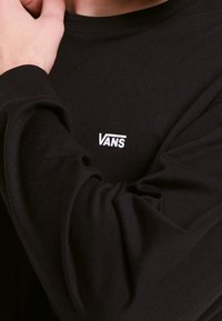 Musta pitkähihainen paita, joka on valmistettu pehmeästä materiaalista ja jossa on valkoinen "VANS"-logo rinnassa. Mukavat ribbineulokset hihansuissa.