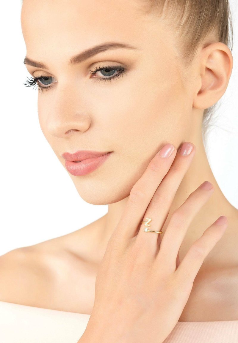 Anello d'oro con un design a zigzag e una piccola pietra preziosa trasparente. Modella che sfiora delicatamente il suo viso, mettendo in mostra l'anello sul suo dito.