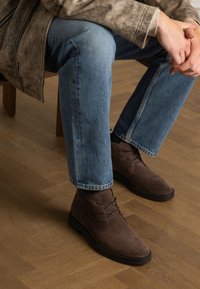 Bottes chukka en suède marron avec un bout arrondi, un laçage et une semelle en caoutchouc. Portées avec un jean bleu et une veste grise texturée.