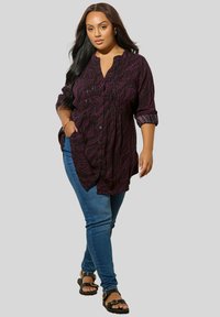 Camicia a bottoni color prugna con una texture a fantasia, caratterizzata da pieghe e un colletto, abbinata a jeans skinny blu e sandali neri.