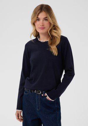 Femme aux longs cheveux blonds ondulés, portant une chemise à manches longues bleu marine et un jean bleu foncé avec une ceinture noire, debout avec une main dans la poche.