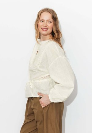 Femme souriante aux longs cheveux portant un chemisier blanc texturé à manches bouffantes et un pantalon marron taille haute, debout devant un fond blanc.