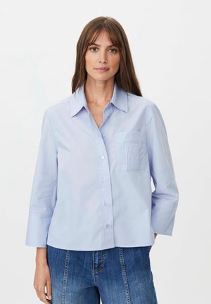 Kvinde med glat brunt hår bærer en lyseblå skjorte med knapper og trekvartærmer samt blå jeans mod en hvid baggrund.