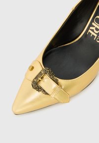 Pointed-toe gold leather shoe with a decorative strap and ornate buckle, featuring a smooth texture and black interior.