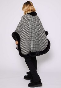 Czarno-biała peleryna w wzór houndstooth z czarnym sztucznym futrem, o swobodnym kroju i szerokich rękawach, noszona na czarnych legginsach.