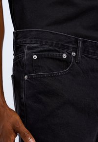 Black denim jeans featuring a classic fit, five-pocket design, silver rivets, and a textured fabric. Hand positioned on the waistband.
