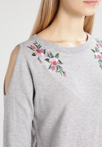 Grå tröja med blommig broderi i rosa, grönt och blått. Har utskurna axlar och en rund halsringning med en mjuk textur.