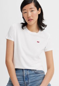 Weißes Baumwoll-T-Shirt mit kurzen Ärmeln, das ein kleines rotes Levi's-Logo auf der Brust trägt. Kombiniert mit blauen Jeans.