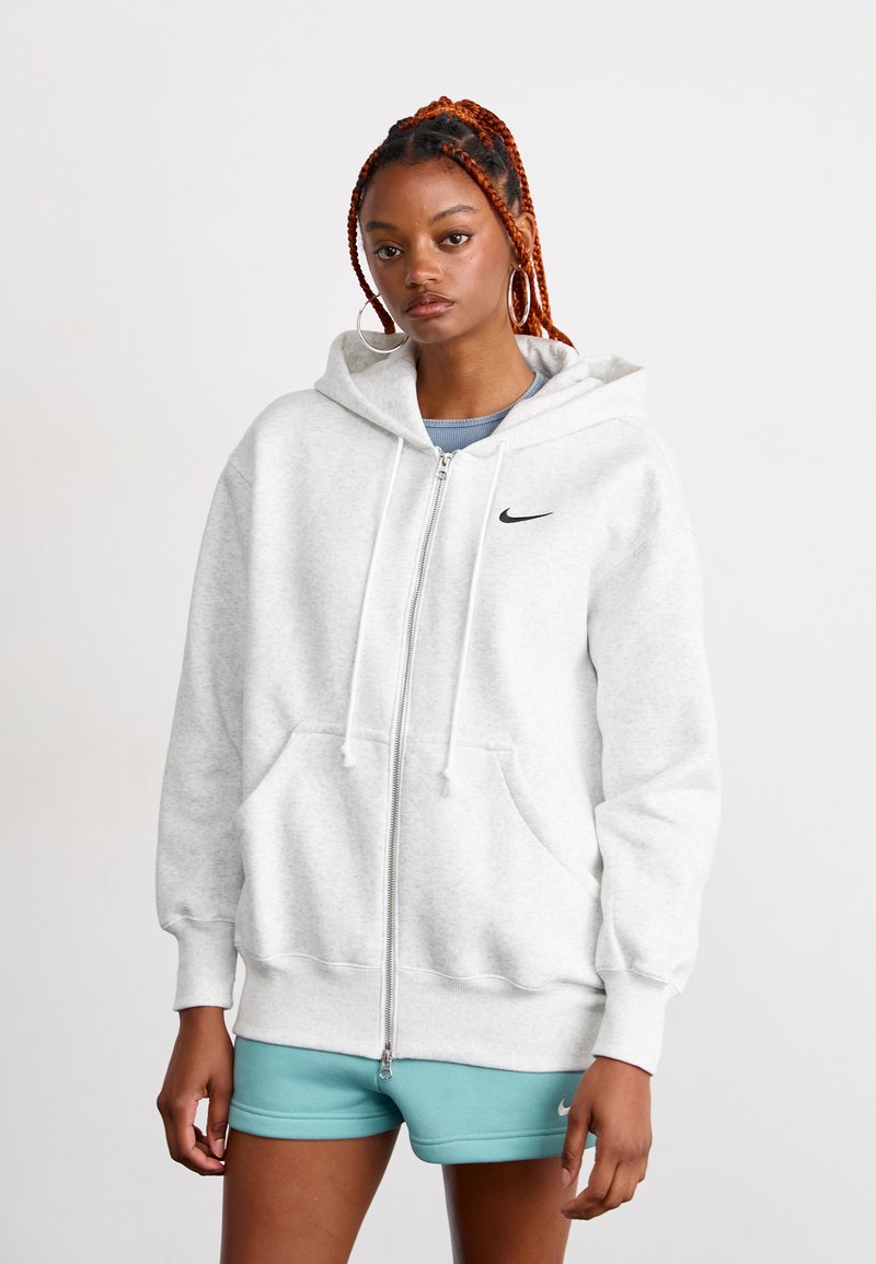 Ung kvinne med flettet rød hår som har på seg store hoops-øredobber, en lys grå hettejakke med glidelås, og teal treningsshorts som står mot en enkel bakgrunn.