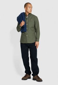 Camicia verde oliva con bottoni, maniche lunghe, abbinata a jeans scuri. Un maglione blu a coste è appoggiato su un braccio. Scarpe marroni con lacci.
