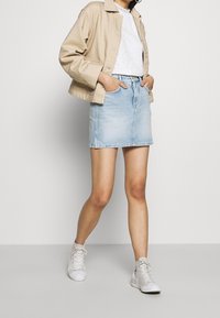 Jupe en denim bleu clair à coupe droite, associée à un haut en dentelle blanche et une veste courte beige ; stylisée avec des baskets blanches montantes.