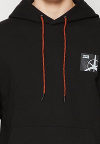 Černá mikina s kapsou na klokany, s červenými stahovacími šňůrkami a malou nášivkou loga na horní levé straně. Texturovaná tkanina.