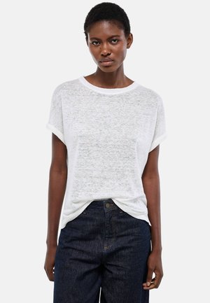 Femme portant un haut blanc à manches courtes amples, rentré dans un jean taille haute bleu foncé, debout devant un fond clair uni.