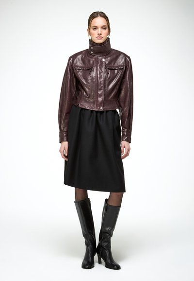 Femme portant une veste en cuir marron foncé, une jupe noire mi-genou, des collants transparents et des bottes à talons noires montant jusqu'aux genoux, debout sur un fond blanc.