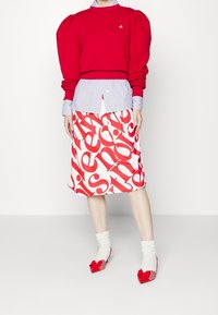 Sudadera roja con mangas abullonadas sobre una camisa de rayas azules, combinada con una falda blanca hasta la rodilla con texto rojo llamativo y zapatos con detalles en rojo.