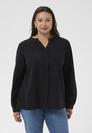 Kobieta o długich ciemnych włosach, ubrana w czarną bluzkę na guziki i niebieskie dżinsy, stojąca na tle jednolitego jasnego tła.