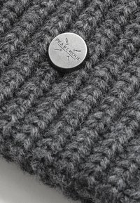 Šedá pletená tkanina s texturovaným vzorem, s kulatým kovovým knoflíkem vyrytým nápisem "PEARL WOOL" a šipkami.