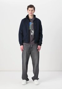 Giacca leggera blu navy con colletto alla coreana, zip frontale, t-shirt nera sotto e jeans grigi a vestibilità ampia, abbinati a sneakers bianche.