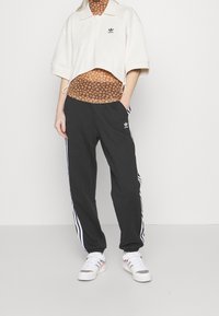 Person wearing cropped white Adidas polo, brown floral sheer top, black Adidas track pants with white stripes, and white sneakers, standing with one hand in pocket.
