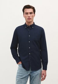 Marineblaues Langarmhemd mit Knöpfen, Brusttasche, weißen Knöpfen und Kragen. Getragen mit hellblauen Jeans. Einfarbiger Hintergrund.