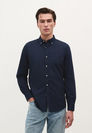 Marineblaues Langarmhemd mit Knöpfen, Brusttasche, weißen Knöpfen und Kragen. Getragen mit hellblauen Jeans. Einfarbiger Hintergrund.