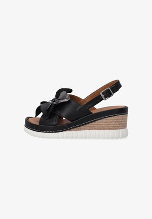 Černé kožené sandály na klínu s dekorativní mašlí a nastavitelným pásem kolem kotníku. Vlastnosti zahrnují texturovanou bílou platformu a dřevěný klínový podpatek.