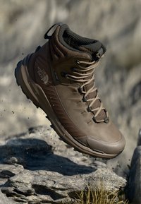 Braune Wanderstiefel mit strukturiertem Obermaterial, gebundenen Schnürsenkeln und gepolstertem Kragen, schwebend über felsigem Terrain mit Staubpartikeln.