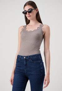 Ribbed brown tank top with scalloped lace trim, paired with high-waisted dark blue jeans. The model stands against a neutral background.