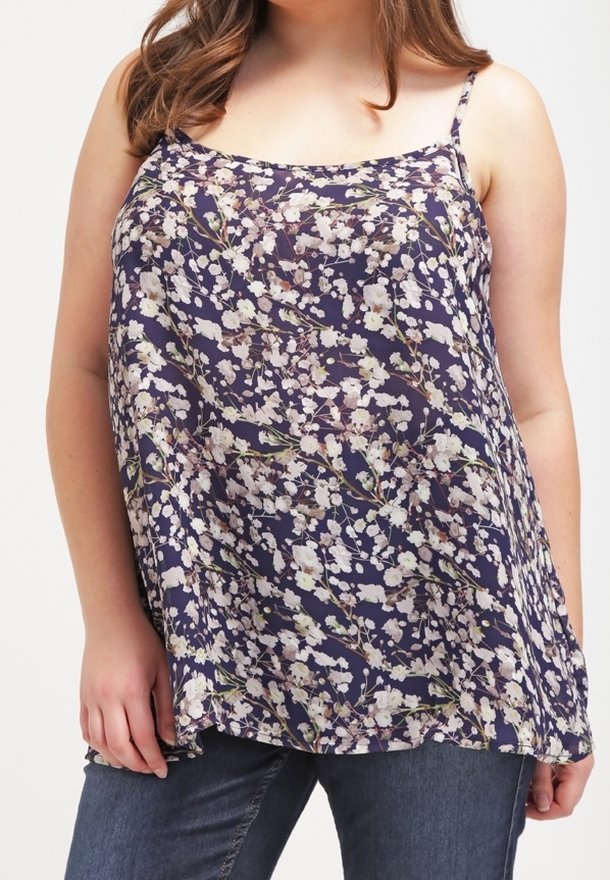 Femme portant un haut sans manches à fleurs bleu marine avec des bretelles fines et un jean bleu foncé, debout devant un fond uni.