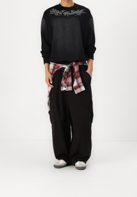 Uomo che indossa una felpa nera con il testo "Eternal Redemption", pantaloni neri larghi, scarpe da ginnastica bianche e una camicia a quadri rossa legata in vita.