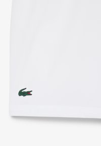 Tessuto bianco con piccolo logo coccodrillo verde ricamato vicino al bordo inferiore sul lato sinistro.