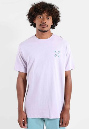 Homme aux cheveux bouclés portant un t-shirt violet clair avec un petit imprimé floral turquoise sur la poitrine et un pantalon bleu clair, debout de manière neutre.