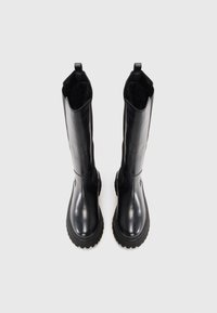 Une paire de bottes en cuir noires montantes jusqu'aux genoux avec des semelles crantées épaisses, debout sur un fond gris clair.