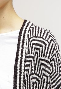 Detailní záběr na rameno osoby, která má na sobě černobílý pletený oděv s texturou a geometrickým vzorem připomínajícím bludiště přes bílou košili.