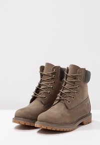 Bruine leren enkelboots met metalen oogjes en olijfgroene veters. Heeft een stevige rubberen zool en een textielkraag voor extra ondersteuning.