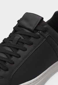 Gros plan sur une basket en cuir noir avec des lacets noirs et une semelle en caoutchouc blanche, montrant les détails des panneaux latéraux cousus et perforés.