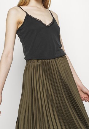 Kvinde iført sort blondekantet camisole og olivengrøn plisseret nederdel, vist fra brystet til midt på låret mod en ensfarvet hvid baggrund.