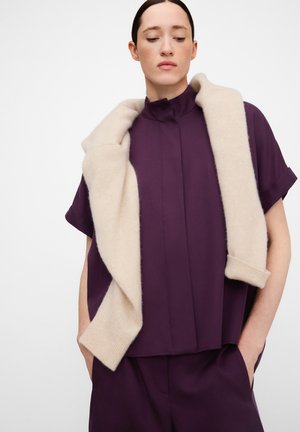 Frau trägt ein dunkelviolettes kurzärmeliges Oberteil und passende Hosen, mit einem cremefarbenen Pullover über den Schultern, die Hände in den Taschen.