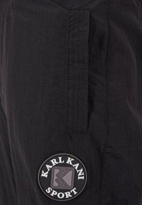 Pantaloni scurti cargo negri cu o patch cu "KARL KANI SPORT." Include buzunare laterale și detalii de cusătură vizibile. Fabricat dintr-un material ușor.