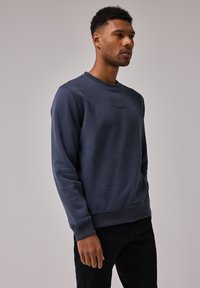 Granatowy sweter z okrągłym dekoltem wykonany z miękkiego materiału, z ściągaczami przy mankietach i u dołu. Posiada małe, nadrukowane logo z przodu.