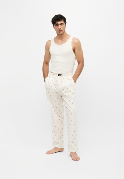 Homme en chemise blanche sans manches et pantalon de pyjama blanc avec un petit motif, debout pieds nus sur un fond blanc uni.