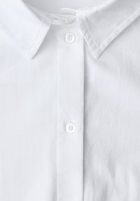 Chemise en coton blanc avec un col classique, dotée d'une fermeture à boutons sur le devant et d'une texture lisse, mettant en avant un design simple et épuré.