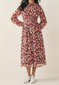 Robe à imprimé floral avec manches longues, taille froncée et encolure en trou de serrure. Couleurs comprenant le rose, le blanc et l'orange sur un fond foncé. Baskets blanches.